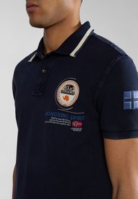 Námořnická polo košile s texturovaným povrchem, s pruhovaným límcem, vyšitými nášivkami a textovým detailem v červené a bílé barvě na přední straně.