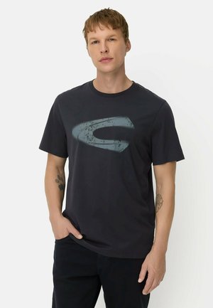 Junger Mann mit kurzen Haaren, der ein schwarzes T-Shirt mit einem großen ausgewaschenen blauen Logo vorne trägt und mit einer Hand in der Tasche steht.