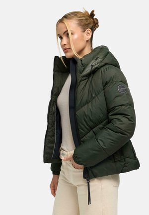 Femme aux cheveux blonds en chignon porte une doudoune vert foncé ouverte sur un haut beige et un pantalon crème, debout devant un fond uni.
