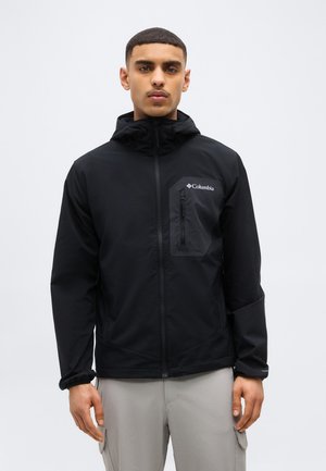 Muž v čiernej bunde Columbia na zips s kapucňou a sivými nohavicami, stojaci pred jednoduchým svetlým pozadím.