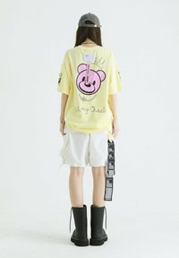 Camiseta amarilla de gran tamaño con un gr�áfico de un oso rosa, combinada con pantalones cortos blancos y botas negras de tobillo; cuenta con etiquetas y diseños de mangas en contraste.