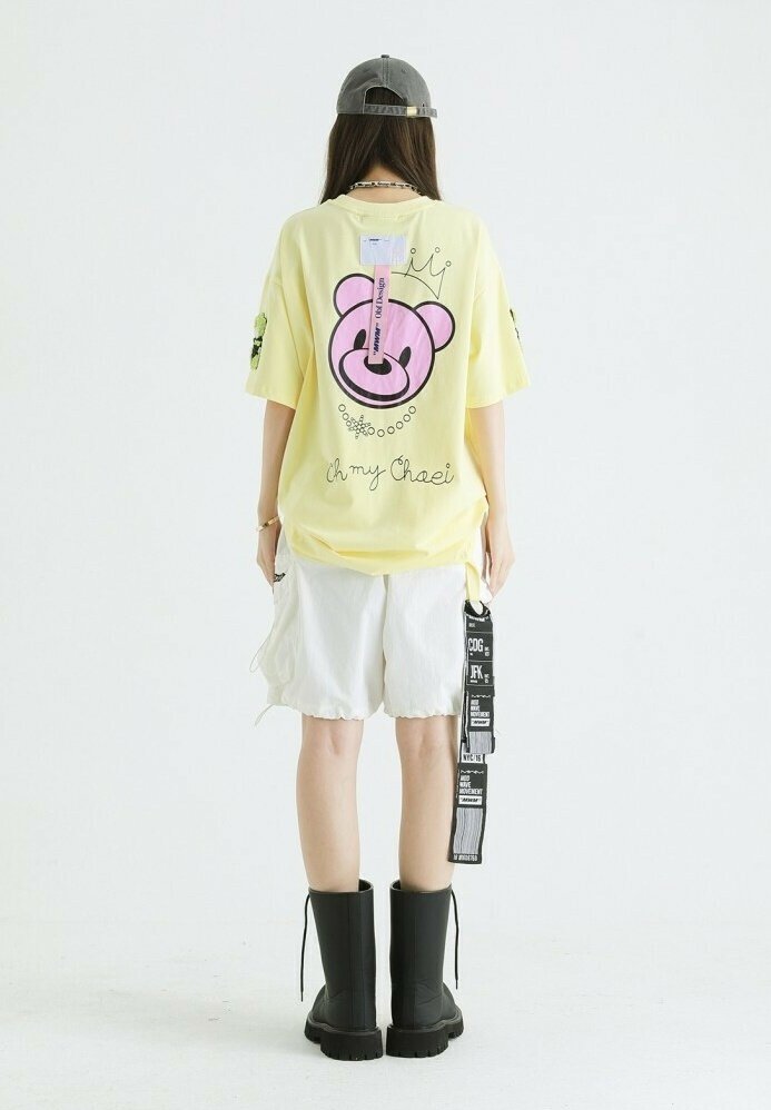 Camiseta amarilla de gran tamaño con un gráfico de un oso rosa, combinada con pantalones cortos blancos y botas negras de tobillo; cuenta con etiquetas y diseños de mangas en contraste.