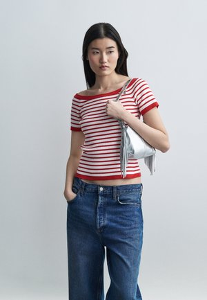 Mlada ženska z dolgimi temnimi lasmi, oblečena v rdečo in belo trakasto majico ter modre kavbojke, drži majhno srebrno torbico z resicami na sivi podlagi.