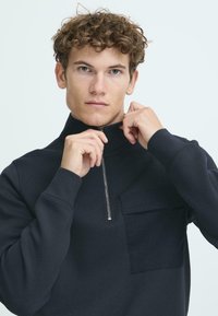 Black pullover with a quarter-zip collar, featuring a front pocket and ribbed cuffs. Smooth fabric with a matte texture.