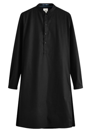 Homme portant une kurta noire à manches longues avec des boutons, un pantalon noir slim et des mocassins noirs, debout sur un sol en bois foncé.