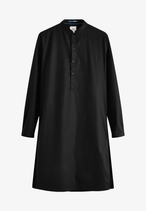 Hombre vestido con una kurta negra de manga larga con botones, pantalones negros ajustados y mocasines negros de pie sobre suelo de madera oscura.