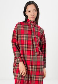 Rot-schwarz kariertes Kleid mit hohem Kragen, weicher Textur, langen Ärmeln und einem Reißverschluss auf der Vorderseite. Mit seitlichen Taschen.