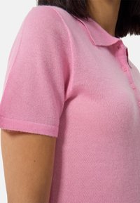 Rosa kortärmad polo-tröja gjord av mjukt tyg med krage och detalj av tre knappblåsnings. Slät textur och jämn färg.