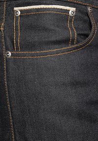 Svarta denimjeans med orange sömmar, synliga nitar och en liten framficka, som visar en slät textur och en figurnära design.