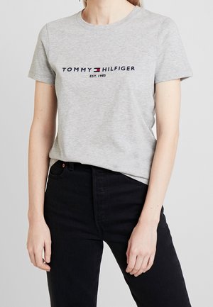 Person iført en lysegrå Tommy Hilfiger T-shirt med logo og sorte højtaljede bukser mod en ensfarvet baggrund.