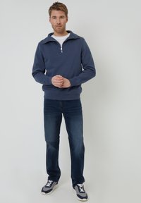 Granatowy sweter z półzamkiem, prążkowane mankiety, zestawiony z ciemnoniebieskimi dżinsami i szarymi trampkami z białymi akcentami. Pod spodem gładka biała koszulka.