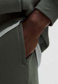 Khaki pulóver bordás mandzsettákkal, cipzáros zsebbel. Fehér aláöltözővel rétegezve. A textúrázott anyag látható.