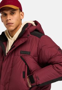 Chaqueta de plumas burdeos con cuello y detalles en negro. Presenta un parche de logo, bolsillos con cremallera y puños ajustables. Textura suave e insulated.