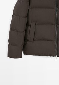 Giacca a vento imbottita con un design trapuntato di colore marrone scuro. Presenta una chiusura frontale con zip e una tasca laterale sul maniche sinistro. Tessuto liscio e morbido.