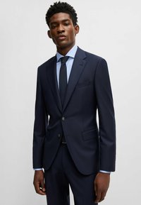 Marineblauer Anzug mit strukturierter Gestaltung, einreihige Jacke, Reverskragen und zwei Knöpfen. Kombiniert mit einem hellblau karierten Hemd und einer dunklen Krawatte.