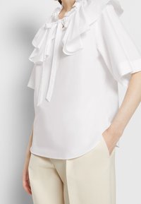 Blouse blanche avec un col à volants oversize, noué au cou. Manches courtes, coupe décontractée, confectionnée en tissu lisse, portée avec un pantalon beige.