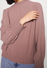 Blusa de manga larga en rosa empolvado con un collar alto, puños fruncidos y dobladillo inferior. Confeccionada en una tela suave y con un corte relajado.