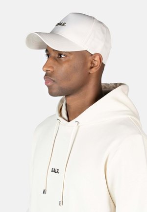 Gorra de béisbol blanca con visera curva, que presenta un logo bordado en negro. La lleva puesta un modelo con una sudadera de color claro con cordones.