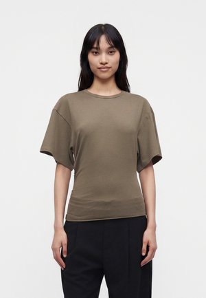 Woman with long dark hair wearing an olive green short-sleeve shirt and black pants, standing against a plain light background.