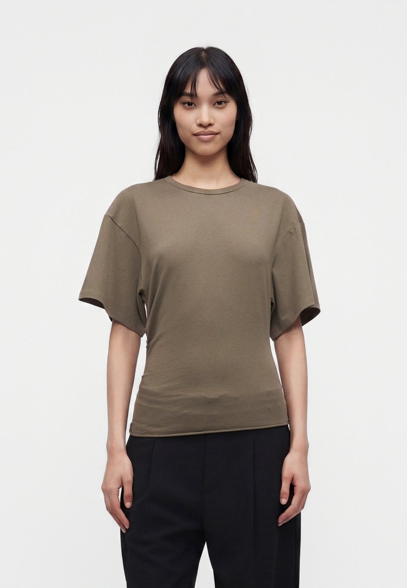Femme aux longs cheveux foncés portant une chemise à manches courtes vert olive et un pantalon noir, debout devant un fond clair uni.
