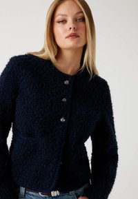 Młoda kobieta z prostymi blond włosami, ubrana w teksturowaną, ciemnoniebieską kurtkę zapinaną na guziki i jasnoniebieskie dżinsy, na tle jasnego, gładkiego tła.