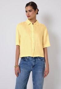 Camicia di lino gialla a maniche corte con bottoni frontali, colletto e design corto. Indossata con jeans in denim blu a vita alta.