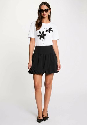 Žena s černými slunečními brýlemi, bílým krátkým topem s černým květinovým vzorem, černou nafouklou mini sukní a černými špičatými podpatky.