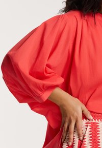 Blusa oversize corallo con maniche a palloncino, caratterizzata da dettagli arricciati sulle spalle. Abbinata a una gonna strutturata con motivo rosso e bianco.
