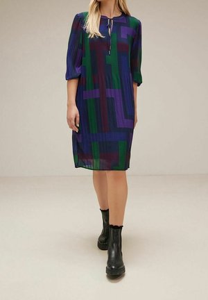 Vrouw draagt een knielange jurk met een multicolor geometrisch patroon, driekwartmouwen en zwarte enkellaarsjes, staand op een effen vloer.