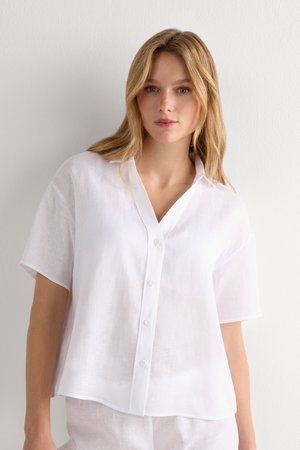 Blond vrouw die een losse, kortemouwen witte linnen blouse met knopen draagt tegen een effen lichte achtergrond.