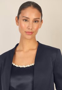 Top di raso blu nave con scollatura accentuata da perle. Vestibilità sartoriale con colletto leggermente aperto, texture liscia e leggera lucentezza.