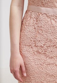 Roze kantjurk met bloemenpatroon, met een gladde textuur en een bijpassende satijnen riem in de taille, gedeeltelijk bedekte hand.