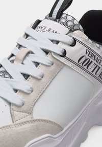 Κοντινό πλάνο λευκού και μπεζ αθλητικού παπουτσιού Versace Couture με χοντρές λευκές κορδόνια και ανάγλυφη λεπτομέρεια στη σόλα.