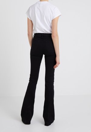 Person trägt ein weißes kurzärmeliges Oberteil und hoch taillierte schwarze Schlaghosen, steht mit dem Rücken zur Kamera vor einem schlichten weißen Hintergrund.