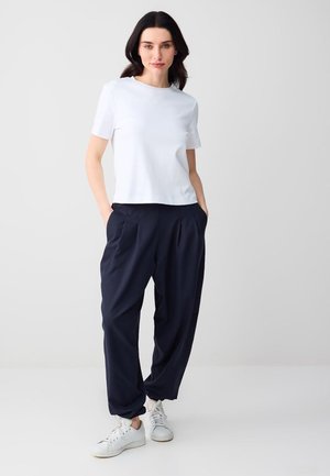 Vrouw staand met handen in zakken, gekleed in een wit T-shirt met korte mouwen, marineblauwe geplooide broek en witte sneakers tegen een effen achtergrond.