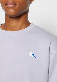 Felpa lilla con scollo rotondo, caratterizzata da un piccolo ricamo di un uccello blu e bianco nella zona sinistra del petto.