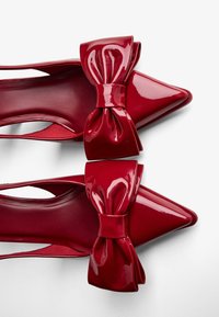 Rdeče lakirane usnjene petke s zašiljenimi prsti in velikim naglavnim poudarkom spredaj. Eleganten dizajn s stranskimi izrezi.