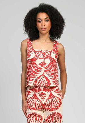 Femme aux cheveux bouclés portant un haut sans manches boutonné à thème squelette rouge et crème et un short assorti, debout avec une main dans la poche.