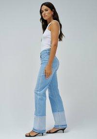 Svetlo modri jeansi s širokimi hlačnicami in značilnim širokim robom z dvobarvnim šivanjem. Obuti z belo majico brez rokavov in črnimi sandali.