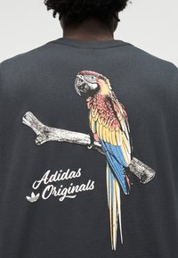 Mørk skjorte med fargerik papegøye sittende på en gren og teksten "Adidas Originals" under fuglen på baksiden.