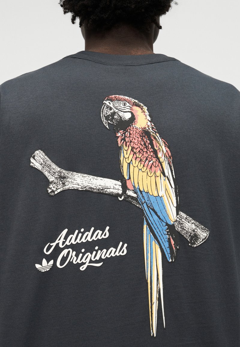 Dunkles Hemd mit buntem Papagei, der auf einem Ast sitzt, und dem Schriftzug "Adidas Originals" unter dem Vogel auf der Rückseite.