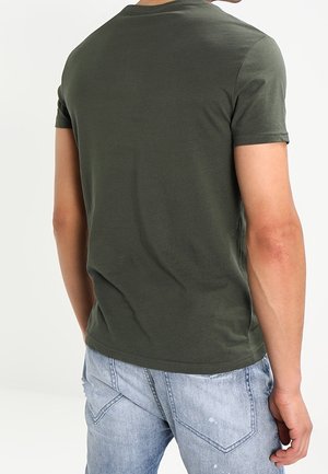 Mann trägt ein olivgrünes kurzärmeliges T-Shirt und hellblaue, abgenutzte Jeans, von hinten vor einem einfarbigen Hintergrund gezeigt.