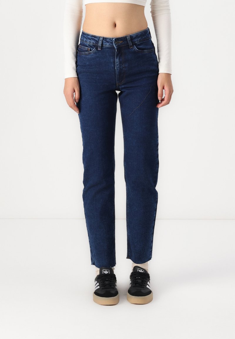 Jeans de mezclilla azul oscuro con un diseño de pierna recta, que presentan una cintura alta, detalles de costura sutiles y un dobladillo deshilachado. Combinados con zapatillas negras.