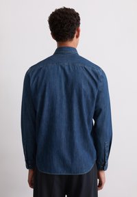 Chemise en denim bleu foncé à manches longues, avec poignets à bouton et col pointu. Présente un ourlet incurvé et un tissu texturé.