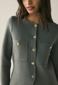 Texturerad grön blazer med gyllene blommönster, med rund halsringning, två framfickor och knappstängning i mitten.
