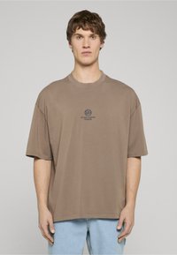 Hnědé oversize bavlněné tričko s kulatým logem a textem uprostřed. Krátké rukávy a kulatý výstřih. Světle modré džíny.