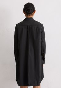 Personne avec des cheveux foncés en chignon portant une robe chemise noire à manches longues, se tenant contre un mur clair uni, de dos face à la caméra.