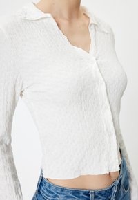 Weißes, strukturiertes Langarmhemd mit Kragen, durchgehender Knopfleiste und leicht verkürztem Saum, kombiniert mit blauen Denim-Jeans.