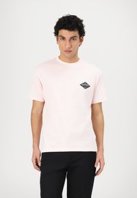 Camiseta de algodón rosa claro con mangas cortas y cuello redondo. Presenta un logotipo de diamante azul oscuro en el área del pecho izquierdo.