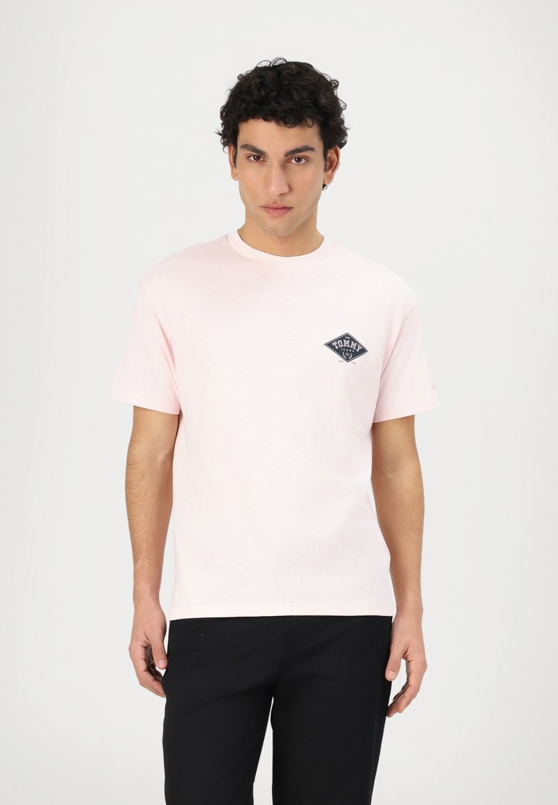 Camiseta de algodón rosa claro con mangas cortas y cuello redondo. Presenta un logotipo de diamante azul oscuro en el área del pecho izquierdo.